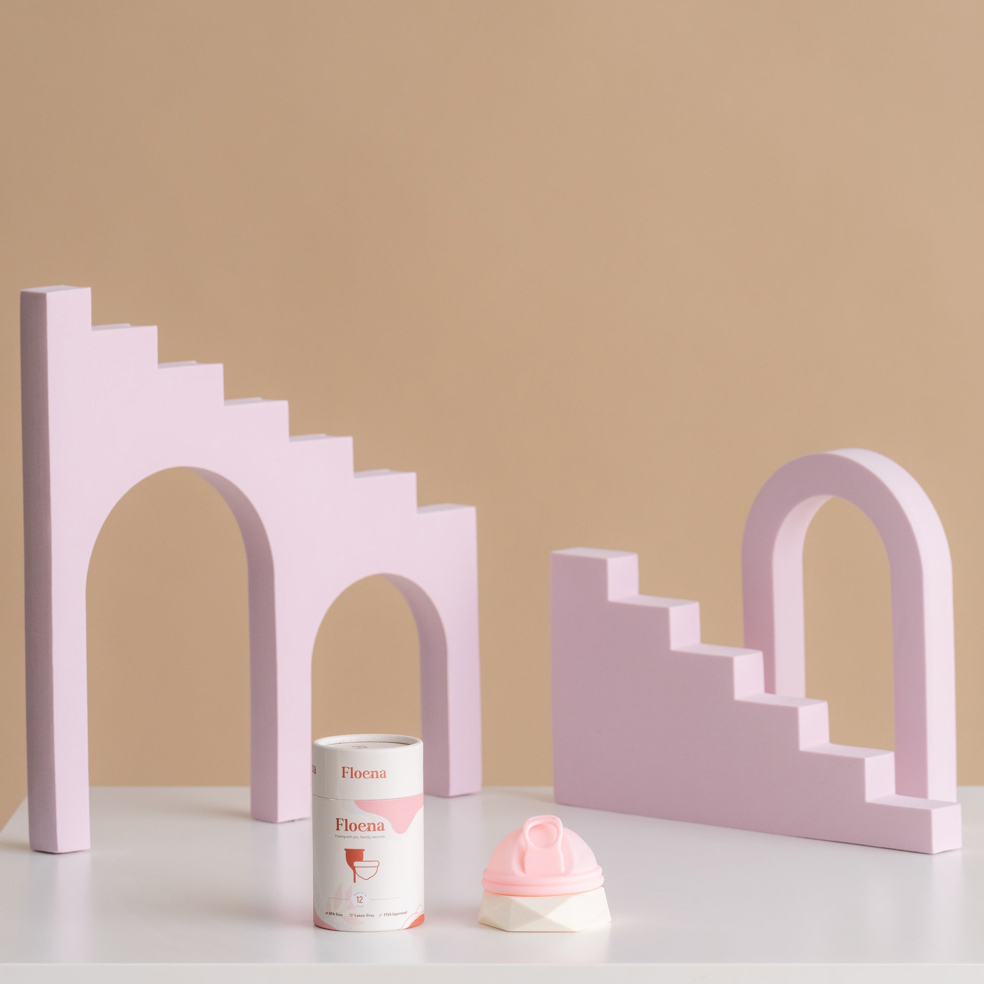 Floena Menstrual Disc — rosa Menstruationsscheibe aus Silikon neben der Verpackung vor verspielten lila geometrischen Blöcken, 12-Stunden-Auslaufschutz