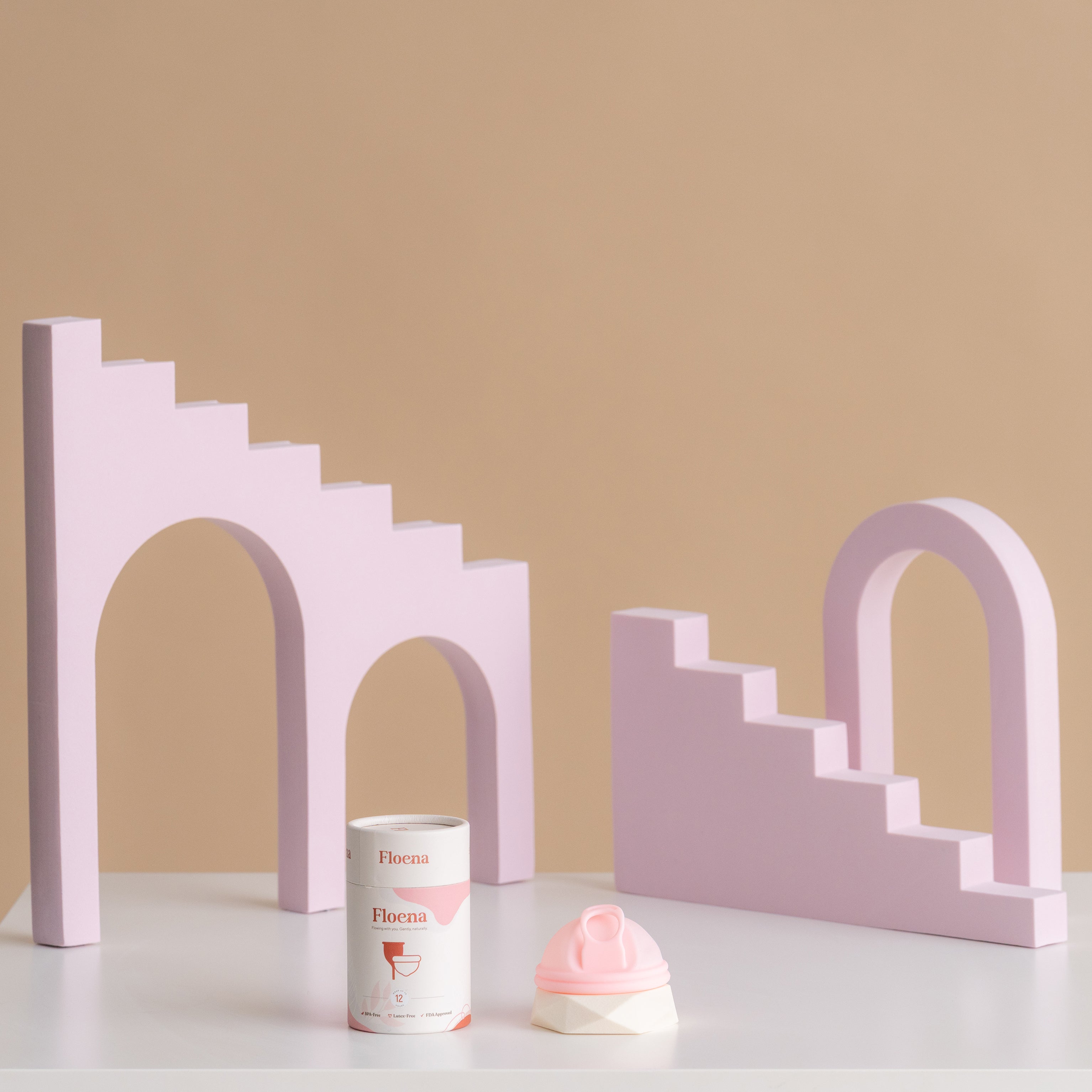Floena Menstrual Disc — rosa Menstruationsscheibe aus Silikon neben der Verpackung vor verspielten lila geometrischen Blöcken, 12-Stunden-Auslaufschutz