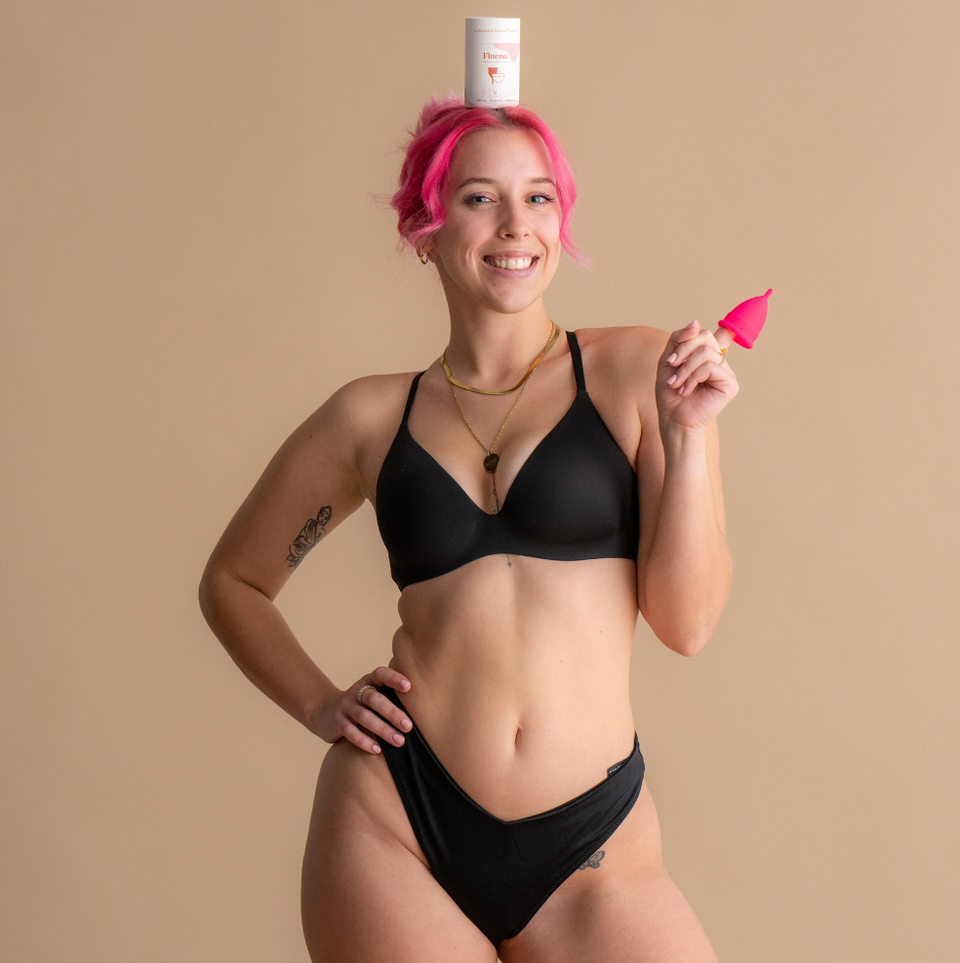 Lächelnde Frau mit pinkfarbenem Haar, die schwarze Unterwäsche trägt und eine leuchtend pinke Floena Ring Cup hält, während ein Floena Produktbehälter auf ihrem Kopf balanciert, fotografiert vor einem weichen neutralen Hintergrund.