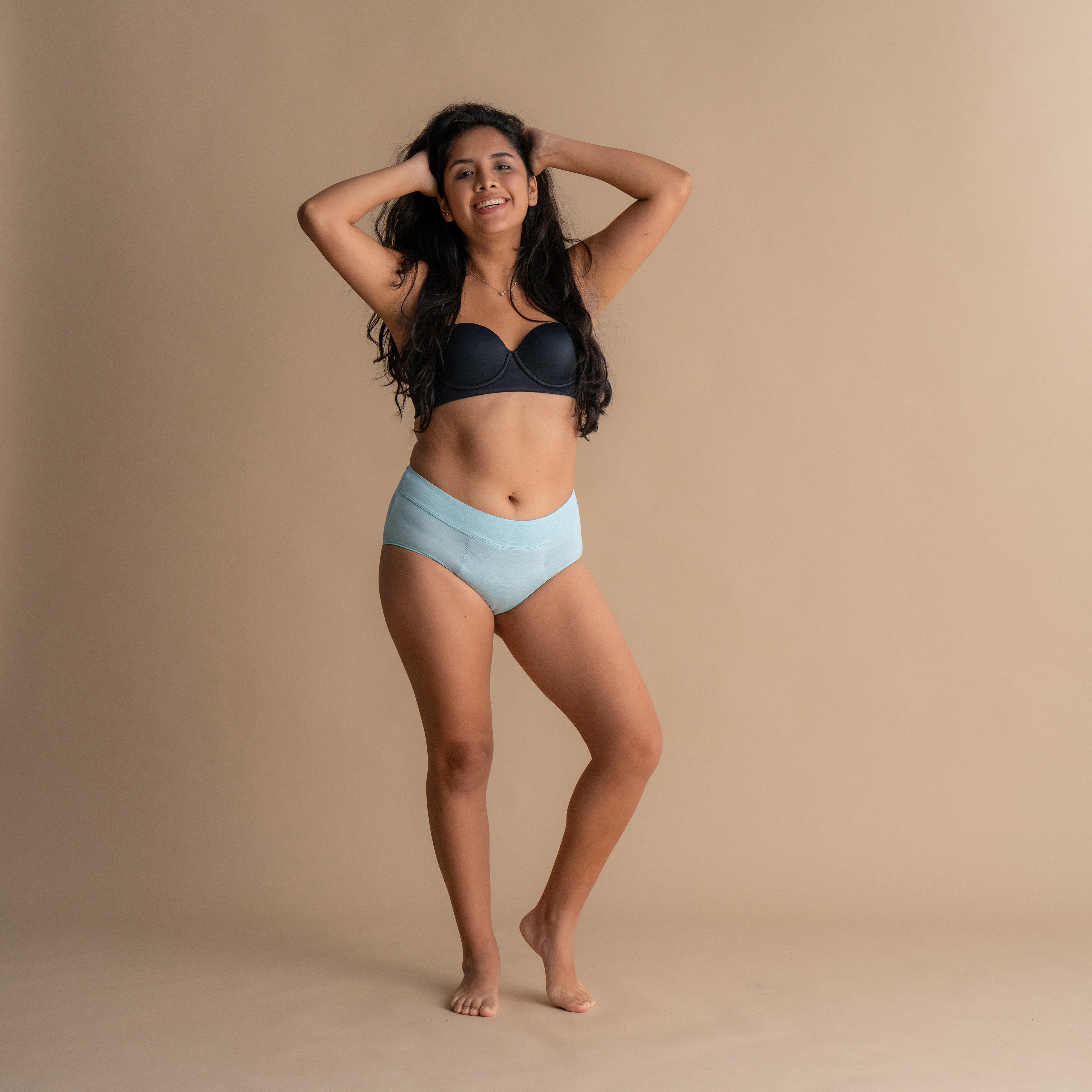 Lächelndes Model in der hellblauen Floena Leakproof Bamboo High Waist Brief, hoch geschnittene Periodenunterwäsche, und schwarzem BH vor beigem Hintergrund
