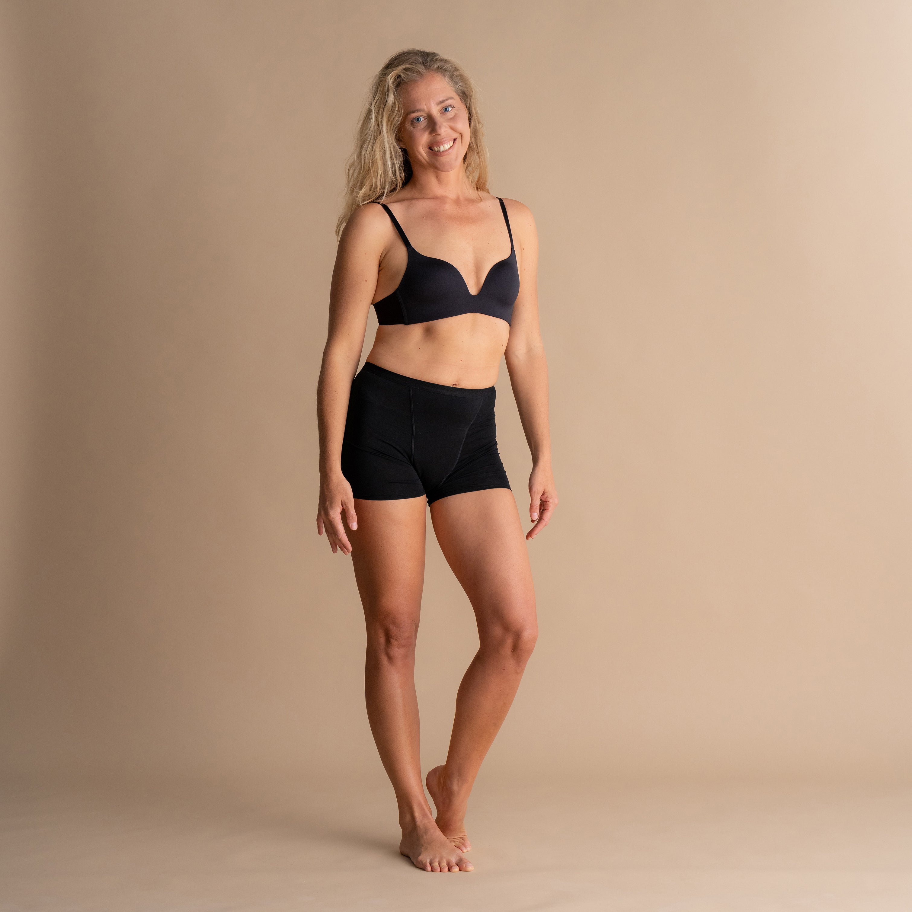 Lächelndes Model in schwarzen Floena Leakproof Bamboo Sleep Short Perioden-Schlafshorts und passendem BH vor beigem Hintergrund