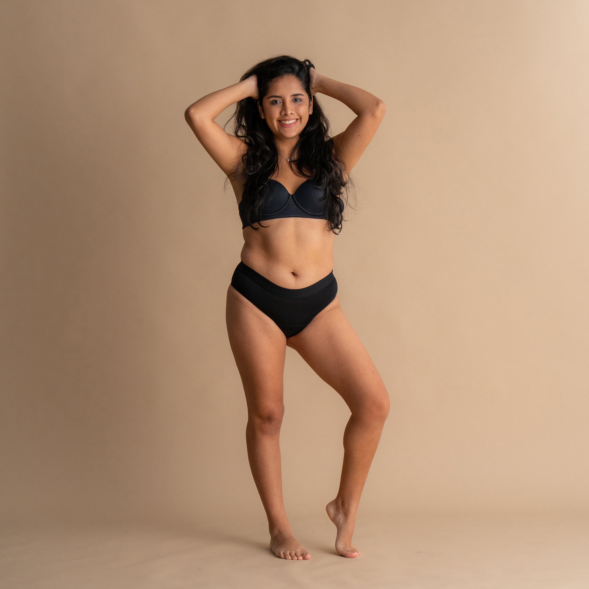Ganzkörper-Frontansicht eines lächelnden Models mit den Händen im Haar in der schwarzen Floena Leakproof Everyday Bamboo Bikini, Periodenunterwäsche aus Bambus