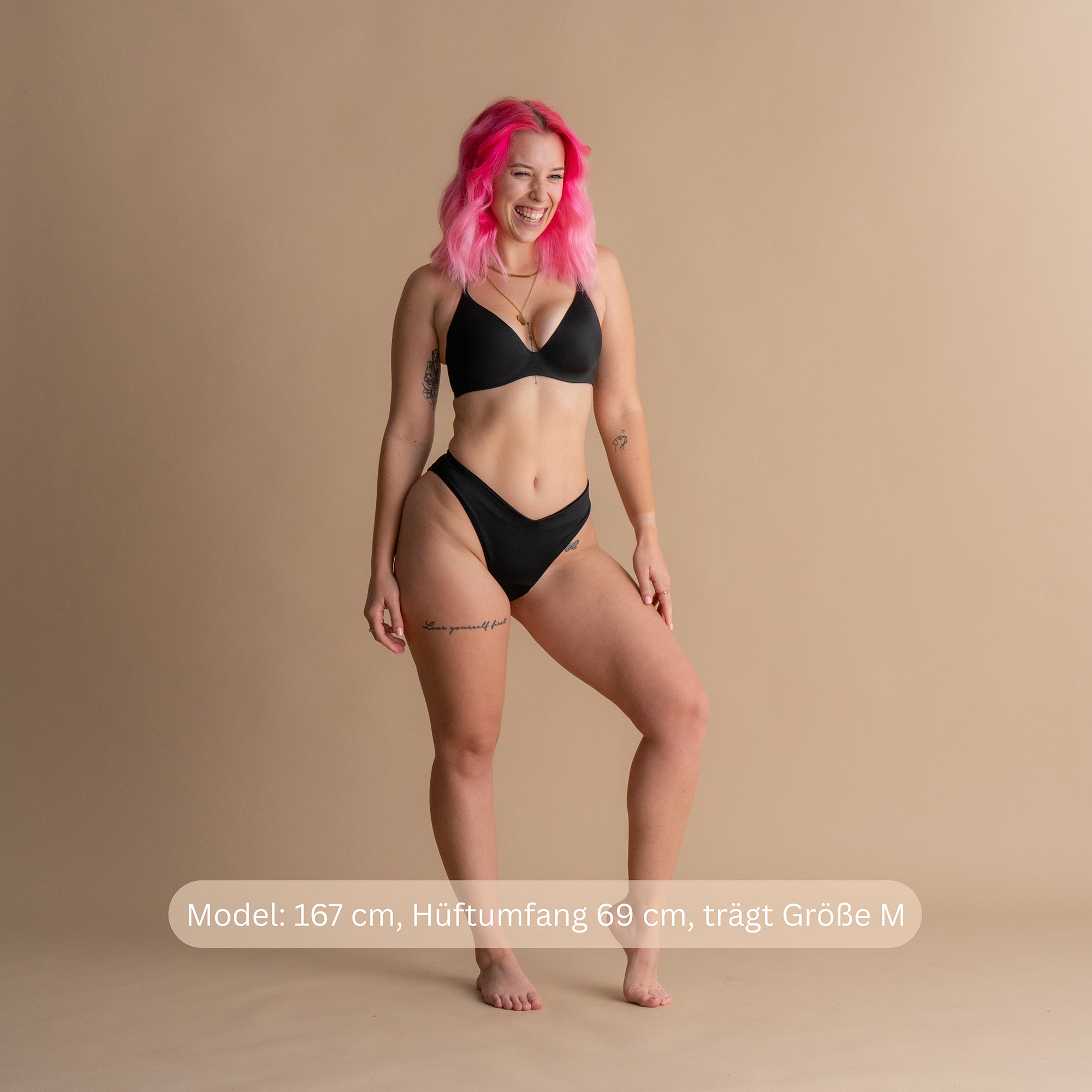 Ganzkörper-Frontansicht eines lächelnden Models mit rosa Haaren in der schwarzen Floena Leakproof Period Swim Bikini Bottom, Periodenbademode, mit passendem Oberteil