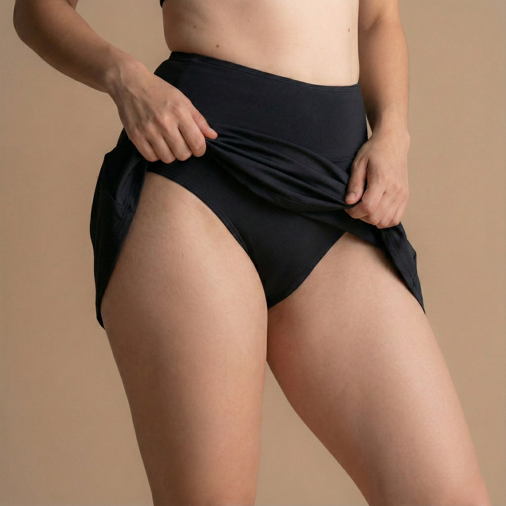 Vordere Seitenansicht eines Models im schwarzen Floena Leakproof Period Swim Skirt, Perioden-Schwimmrock, mit hohem Bund und wasserabweisendem Stoff