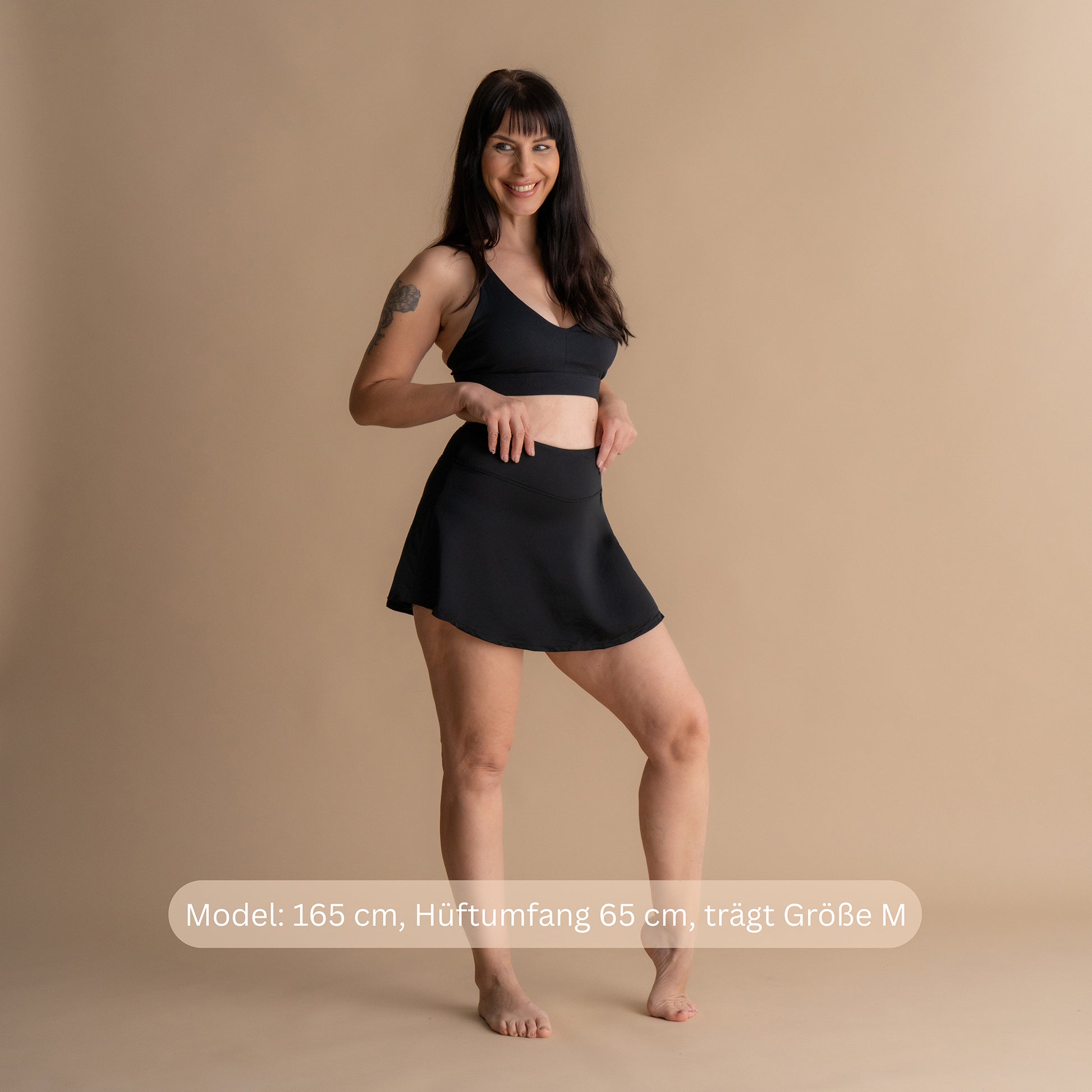 Vorderansicht eines lächelnden Models mit den Händen in den Hüften im schwarzen Floena Leakproof Period Swim Skirt, Perioden-Schwimmrock, mit passendem Bikini-Oberteil