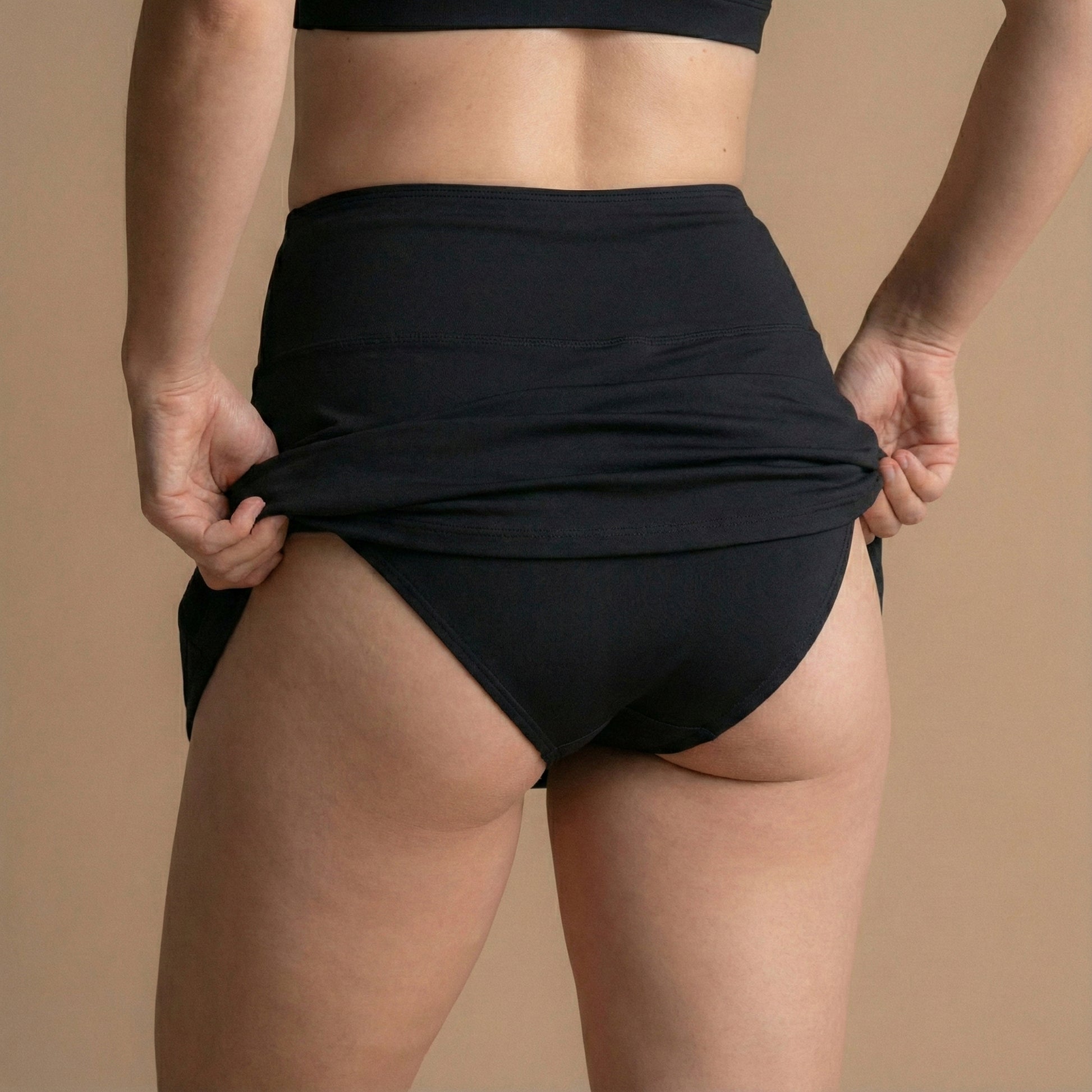 Rückansicht eines Models, das den schwarzen Floena Leakproof Period Swim Skirt, Perioden-Schwimmrock, anpasst, mit voller Abdeckung und hohem Bund