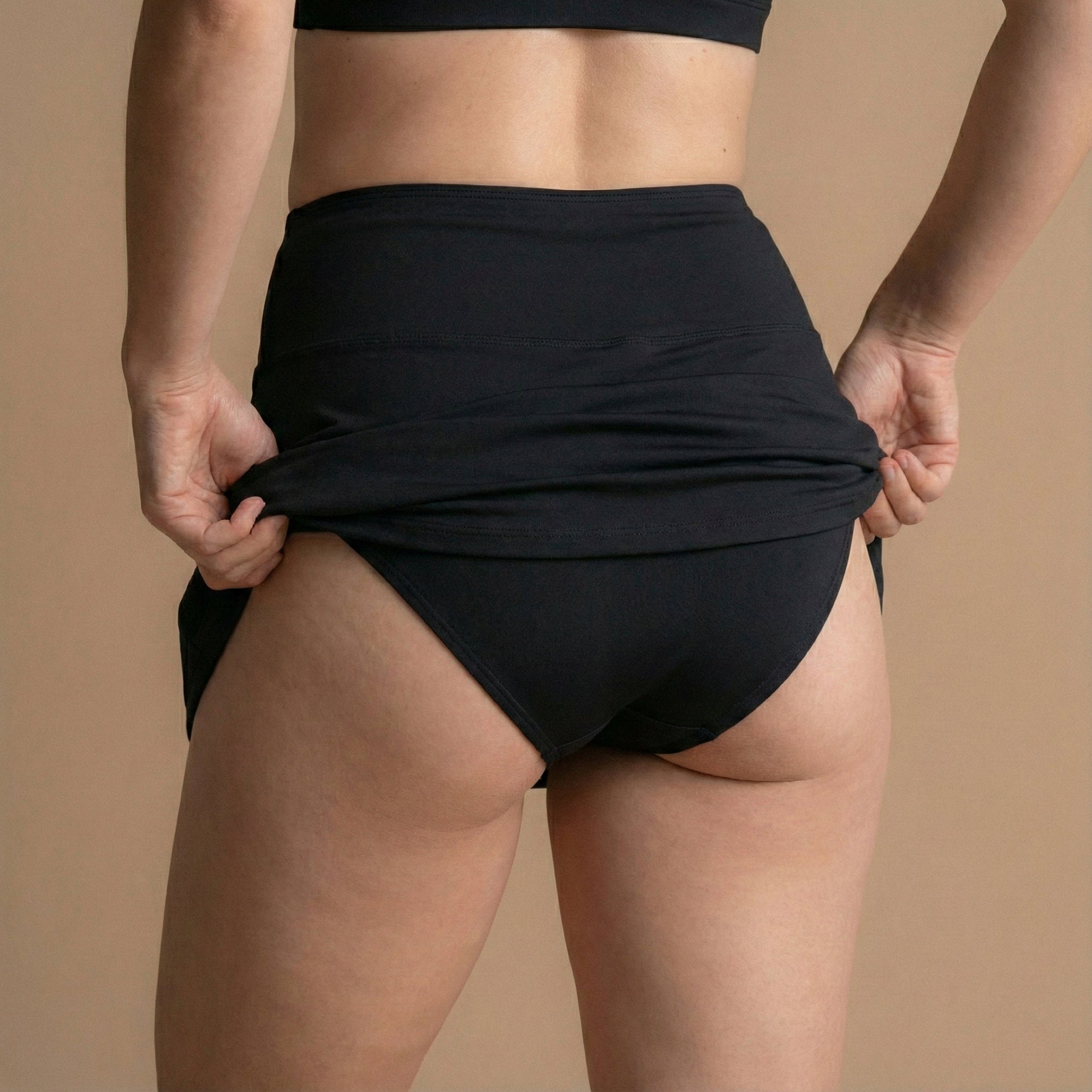 Rückansicht eines Models, das den schwarzen Floena Leakproof Period Swim Skirt, Perioden-Schwimmrock, anpasst, mit voller Abdeckung und hohem Bund