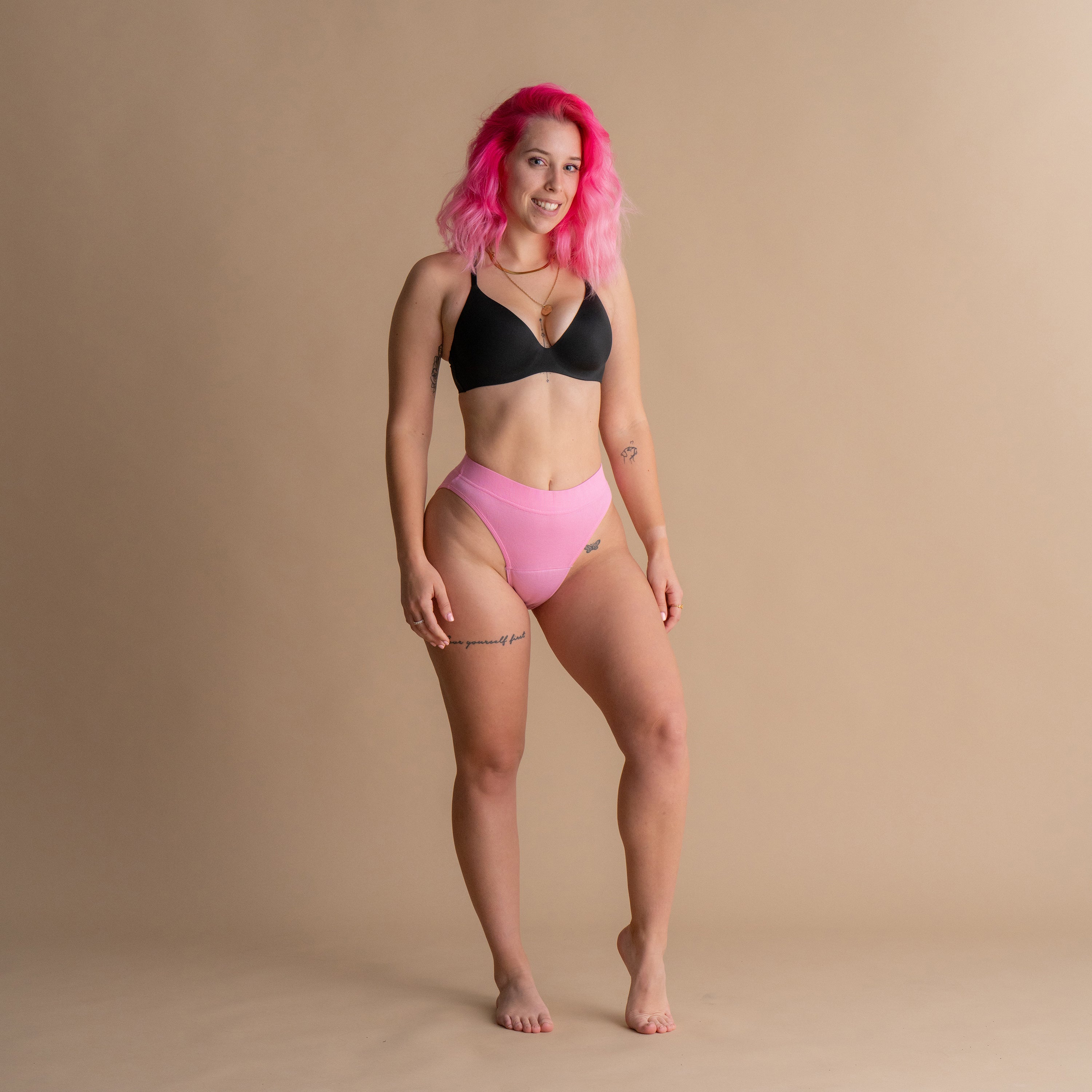 Ganzkörper-Frontansicht eines Models mit rosa Haaren in der rosa Floena Leakproof Ribbed High Waist Brief, gerippte hoch geschnittene Periodenunterwäsche, und schwarzem BH vor beigem Hintergrund
