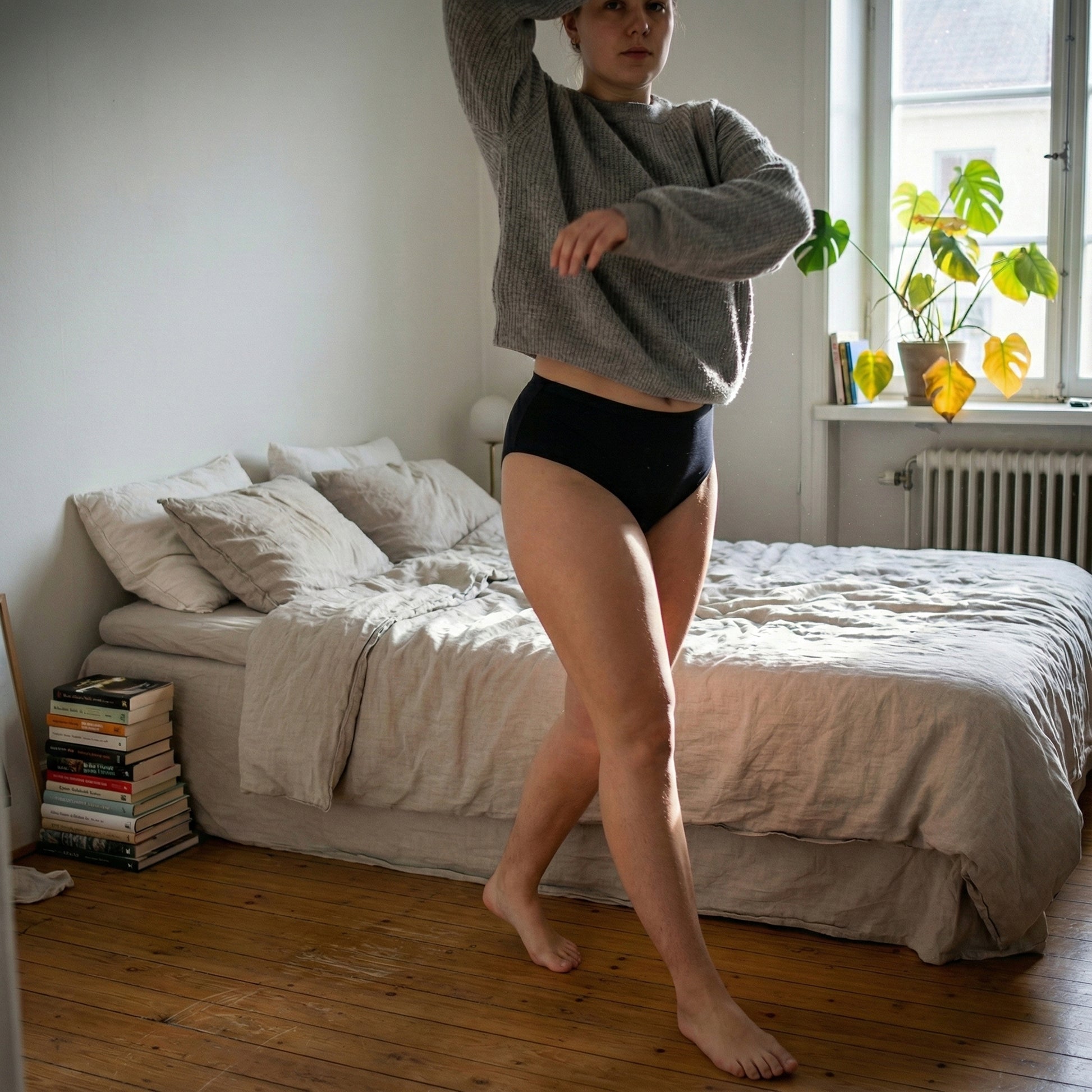 Model in grauem Pullover und der schwarzen Floena Leakproof Seamless High Waist Brief, nahtlose hoch geschnittene Periodenunterwäsche, geht durch ein sonnendurchflutetes Schlafzimmer