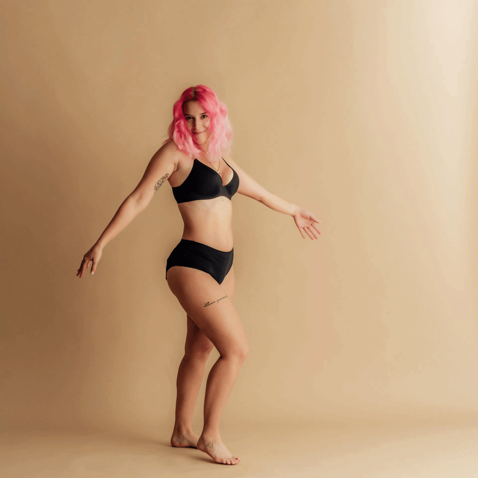 Frau mit pinken Haaren, trägt schwarze Leakproof Period Underwear und tanzt fröhlich in einem Studio, drückt Selbstvertrauen, Komfort und Körperpositivität mit einer spielerischen Pose aus.